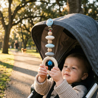 Kind sitzt im Kinderwagen im Freien und hält einen Wagenhänger in Natur- und Blautönen mit Holzperlen, Ring und kleiner integrierter Glocke (ca. 20 cm lang); der Anhänger ist am Verdeck befestigt, im Hintergrund ein sonniger Parkweg mit Bäumen.