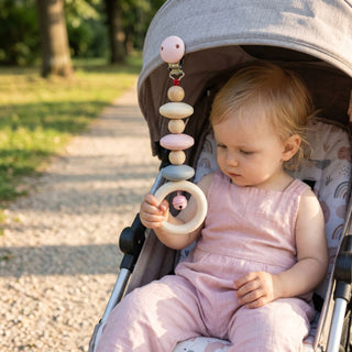 Kind sitzt im Kinderwagen im Freien und greift nach einem Wagenhänger in Natur- und Rosatönen mit Holzperlen, Ring und kleiner Glocke (ca. 20 cm lang), der am Verdeck befestigt ist; im Hintergrund ein sonniger Parkweg mit Bäumen.