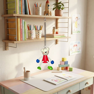Farbig gestaltete Schwingfigur „Bär“, ca. 11 cm groß, aus Holz mit beweglichen Armen und Beinen, an einer Feder unter einem Wandregal aufgehängt. Die Figur hängt über einem Kinderschreibtisch mit Papier, Stiften und Bauklötzen; im Hintergrund befinden sich Bücher, Dekoration und ein Regal.