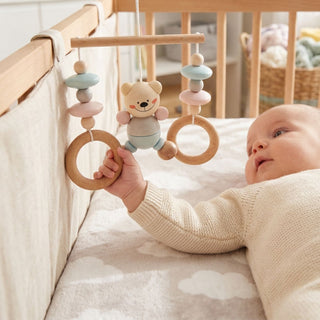 Baby liegt im Kinderbett und greift nach einem hängenden Minitrapez aus Holz mit Bär-Motiv in Natur- und Blautönen; das Spielzeug besteht aus Perlen, Ringen und einer Figur und ist am Bettgestell befestigt, im Hintergrund ein liebevoll eingerichtetes Kinderzimmer.