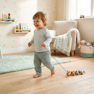 Farbig gestaltete Nachziehraupe „Ida“ aus Holz mit mehreren bunten Kugel-Elementen. Ein kleines Kind läuft barfuß durch ein helles Kinderzimmer und zieht die Raupe an einer Schnur hinter sich her; im Hintergrund sind ein Sessel, Spielzeug und ein Fenster zu sehen.