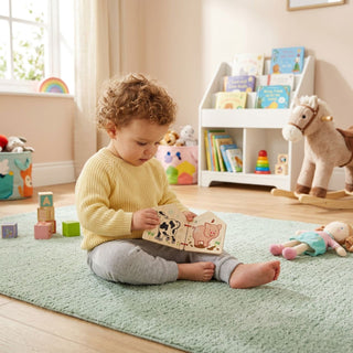 Farbig bedrucktes Bilderbuch „Lustige Tiere“ mit Tiermotiven. Ein kleines Kind sitzt auf einem Teppich im Kinderzimmer und betrachtet das aufgeklappte Buch; im Hintergrund sind Spielzeug, ein Regal mit Kinderbüchern und ein Fenster zu sehen.