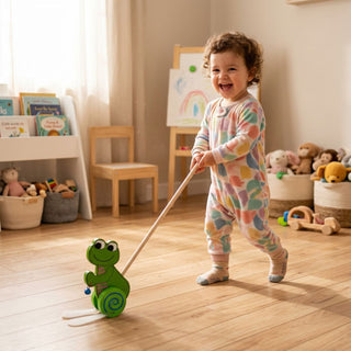 Kleines Kind steht auf Holzboden in einem hellen Raum und schiebt einen grünen, bunten Frosch als Holzspielzeug mit Stab vor sich her; im Hintergrund befinden sich Spielzeugkisten, Bücher und eine Kinderstaffelei.