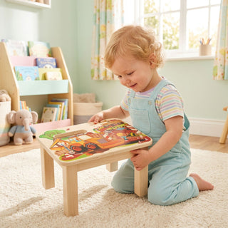 Farbig gestaltete Fußbank „Baustelle“, ca. 17,5 cm hoch, mit Motiv von Baufahrzeugen. Ein kleines Kind kniet auf einem Teppich im Kinderzimmer und betrachtet die bemalte Oberfläche der niedrigen Holz-Fußbank. Im Hintergrund stehen ein Regal mit Kinderbüchern, ein Fenster mit Vorhängen sowie Spielzeug.
