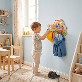 Garderobe „Baustelle“, ca. 37 cm breit, farbig gestaltet mit Motiven von Baufahrzeugen. Ein Kind hängt eine Jacke und einen gelben Bauhelm an die Haken in einem hellen Kinderzimmer; im Hintergrund sind ein Fenster, Möbel und Spielzeug zu sehen.