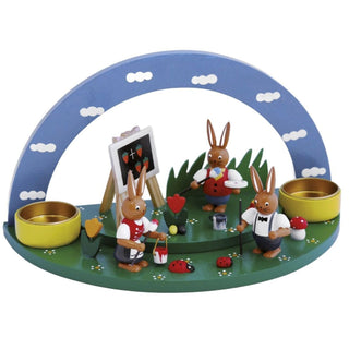 Grüne Blumenwiese mit blauen Himmelsbogen, 2 gelbfarbenen Teelichthaltern und einer farbigen Hasenschule, bestehend aus 3 süßen Osterhasen und kleinen frühlingshaften Details