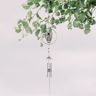 Filigranes Windspiel aus Metall mit stilisierter Eule in einem runden Rahmen, aufgehängt an einer Kette zwischen grünen Zweigen. Unter der Eule hängen silberfarbene Klangstäbe und ein kleiner Kristallanhänger. Der helle, ruhige Hintergrund betont die natürliche und dekorative Gartenszene.