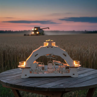 Kerzenhalter für 3 Teelichte „Agrar‑Motiv“, 23 cm, aus naturbelassenem Holz mit detailreich gelaserten Motiven wie Traktor, LKW, Feldmaschinen und Milchkannen, auf einem Holztisch im Freien platziert, vor einem Getreidefeld bei Sonnenuntergang mit einem Mähdrescher im Hintergrund und warm leuchtenden Teelichtern.
