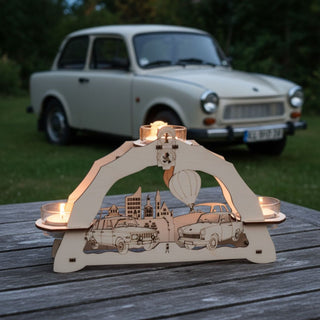 Kerzenhalter für 3 Teelichte „Trabant und Wartburg“, 23 cm, aus naturbelassenem Holz mit filigran gelaserten Motiven zweier klassischer Fahrzeuge sowie Stadtansicht und Heißluftballon, auf einem Holztisch im Freien platziert, im Hintergrund ein Trabant, umgeben von Bäumen, mit warm leuchtenden Teelichtern bei Dämmerung.