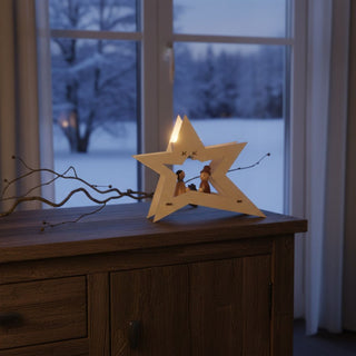 Kerzenhalter für 1 Teelicht „Christi Geburt“, 15 cm, aus naturbelassenem Holz in moderner Sternform mit kleiner Krippenszene im Inneren, dekorativ auf einem Holzsideboard neben Zweigen mit Beeren platziert, vor einem Fenster mit verschneiter Winterlandschaft und warm leuchtendem Kerzenlicht.
