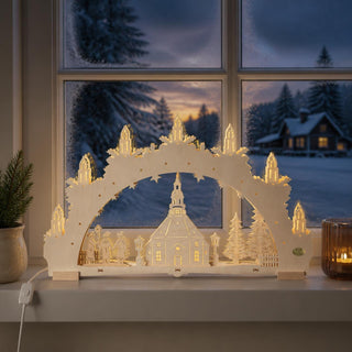 LED-Schwibbogen „Seiffener Kirche“ aus naturfarbenem Holz mit filigraner 3D-Gestaltung. Im Zentrum steht die berühmte Seiffener Rundkirche, umgeben von Tannenbäumen, Laternen und zwei traditionellen Holzfiguren. Sieben kerzenförmige LED-Lichter auf dem Bogen spenden warmweißes Licht und sorgen für eine stimmungsvolle Tiefenwirkung. Der 52 cm breite Schwibbogen steht auf einer Fensterbank vor einer verschneiten Winterlandschaft in der Abenddämmerung und schafft eine festliche Weihnachtsatmosphäre.
