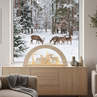 LED Schwibbogen aus naturfarbenem Holz mit filigranem Waldmotiv. Im Inneren des 52 cm breiten Bogens sind mehrere Tannenbäume sowie Hirsche und Rehe dargestellt, die von warmweißen LEDs stimmungsvoll beleuchtet werden. Der Schwibbogen steht auf einer Holzkommode vor einem großen Fenster mit verschneiter Winterlandschaft und äsendem Wild im Hintergrund. Die natürliche Holzoptik und das warme Licht schaffen eine gemütliche, weihnachtliche Atmosphäre.
