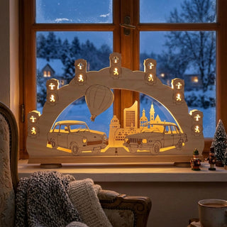 LED-Schwibbogen „(N)ostalgie Trabant und Wartburg“ aus naturbelassenem Holz mit warmweißer LED-Beleuchtung. Das detailreiche Motiv zeigt die legendären DDR-Fahrzeuge Trabant und Wartburg vor einer stilisierten Stadtsilhouette mit Fernsehturm und Heißluftballon. Entlang des Bogens sind beleuchtete Ampelsymbole der bekannten DDR-Ampelmännchen integriert, die dem Design einen besonderen Ostalgie-Charakter verleihen. Der 52 cm breite Schwibbogen steht auf einer Fensterbank vor einer verschneiten Landschaft.