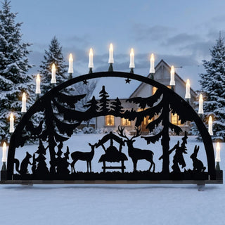 Metallschwibbogen für den Außenbereich „Wald“ in Schwarz mit filigraner Waldszene. Der 150 cm breite Schwibbogen zeigt Rehe an einer Futterkrippe, umgeben von Tannenbäumen und weiteren Waldtieren. Mehrere warm leuchtende Kerzen entlang des Bogens setzen die detailreichen Silhouetten stimmungsvoll in Szene und sorgen für eine festliche Atmosphäre im Außenbereich.