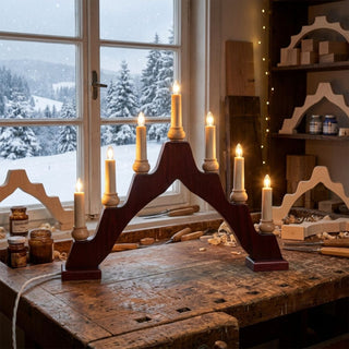 Schwibbogen im Stil eines klassischen Schwedenleuchters aus rot gebeiztem Holz mit sieben warm leuchtenden Kerzen. Der 43 cm breite Lichterbogen steht auf einer rustikalen Werkbank vor einem Fenster mit verschneiter Winterlandschaft. Die Kombination aus dunkelrotem Holz, naturfarbenen Kerzenhaltern und warmem Licht sorgt für eine traditionelle, gemütliche Weihnachtsatmosphäre.