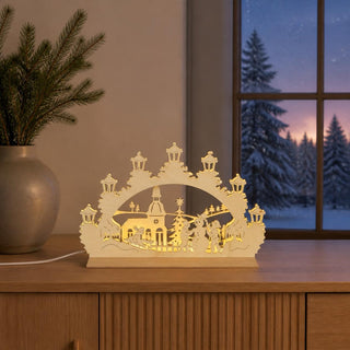 LED-Schwibbogen Mini „Schneemann“ aus naturfarbenem Holz mit winterlicher Dorfszene. Das Motiv zeigt Kinder beim Bauen eines Schneemanns vor einer Kirche und einem geschmückten Weihnachtsbaum. Sieben laternenförmige LED-Lichter sorgen für warmes, stimmungsvolles Licht. Der 24 cm breite Mini-Schwibbogen ist eine festliche Dekoration für Fensterbank, Regal oder Tisch.