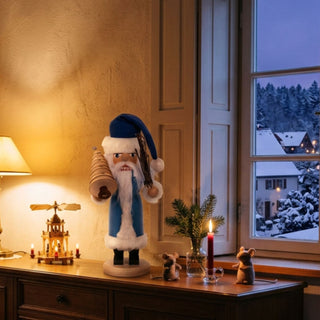 Nussknacker „Weihnachtsmann“ (36 cm) in Blau mit Tannenbaum, stehend auf einem Holzsockel, dekorativ auf einer Kommode mit Kerzen und winterlicher Dorfkulisse vor dem Fenster platziert.