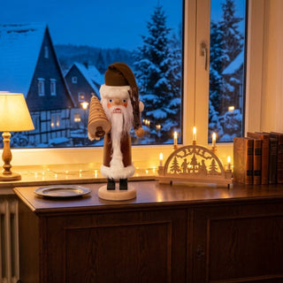 Nussknacker „Weihnachtsmann“ (36 cm) in Braun mit Tannenbaum, stehend auf einem Holzsockel, dekorativ auf einer Kommode neben Schwibbogen und Kerzen vor winterlicher Fensterkulisse platziert.