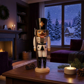Nussknacker „Soldat“ in weißer Uniform (29 cm) mit schwarzem Hut und Gewehr, steht auf einem Holztisch. Umgeben von Kerze, Tannenzweig und Zapfen, vor einem gemütlichen Wohnzimmer mit Kamin und verschneiter Winterlandschaft am Fenster.