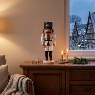 Nussknacker „König“ (36 cm) in Weiß mit Krone und Zepter, stehend auf einem Holzsockel, dekorativ auf einer Kommode mit Kerzen und Winterlandschaft vor dem Fenster platziert.Geben Sie Ihr Feedback zu BizChat ab.