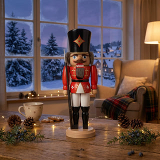 Nussknacker „Soldat“ in roter Uniform (29 cm) mit schwarzem Hut und Gewehr, steht auf einem Holztisch. Umgeben von Lichterkette, Zapfen, Tasse und Gebäck, vor einem gemütlichen Wohnzimmer mit Sessel und verschneiter Winterlandschaft am Fenster.