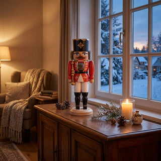 Nussknacker „König“ (36 cm) in Rot mit Krone und Zepter, stehend auf einem Holzsockel, dekorativ auf einer Kommode mit Kerze und winterlicher Fensterlandschaft im Hintergrund platziert.