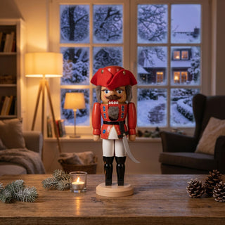 Nussknacker „General“ in roter Uniform (29 cm) mit Hut und Säbel, auf einem Holztisch mit Kerze und Tannenzweigen vor winterlichem Fenster im gemütlichen Wohnzimmer.