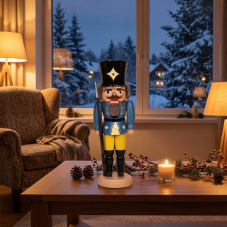 Nussknacker „Soldat“ in blauer Uniform (29 cm) mit schwarzem Hut und Gewehr, steht auf einem Holztisch. Umgeben von Kerze, Zapfen und Beerenzweigen, vor einem gemütlich eingerichteten Wohnzimmer mit Sessel und verschneiter Winterlandschaft am Fenster.