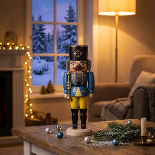 Nussknacker „König“ in blauer Uniform (29 cm) auf einem Tisch, mit Weihnachtsdeko und gemütlichem Wohnzimmer vor winterlichem Fenster.