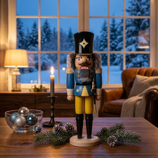 Nussknacker „Husar“ (36 cm) in Blau mit Säbel, stehend auf einem Holzsockel, dekorativ auf einem Tisch mit Kerze, Tannenzweigen und Kugeln vor großer Fensterfront mit winterlicher Landschaft platziert.