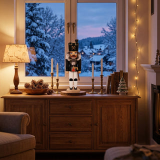 Nussknacker „Bergmann“ (36 cm) in Schwarz mit Axt, stehend auf einem Holzsockel, dekorativ auf einer Kommode mit Kerzen, Büchern und Lichterkette vor winterlicher Fensterlandschaft platziert.