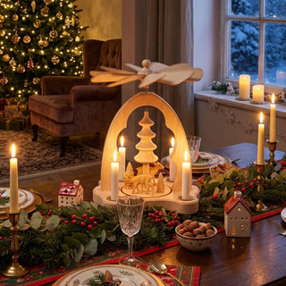 Weihnachtspyramide „Rehe“ aus naturbelassenem Holz (21 cm) für vier Pyramidenkerzen mit Flügelrad. Szene mit Rehen und stilisiertem Tannenbaum unter einem geschwungenen Bogen. Auf einem festlich gedeckten Tisch mit Kerzen, Tannenzweigen und Weihnachtsdekoration vor geschmücktem Weihnachtsbaum und Fenster.
