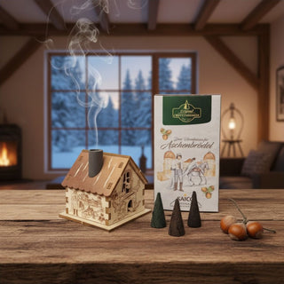 Räuchermännchen „Räucherhaus – Drei Haselnüsse für Aschenbrödel®“ (7 cm), naturbelassen gestaltet als kleines Häuschen mit filigranen Märchenmotiven, inkl. Räucherkerzen, rauchend auf einem Tisch vor gemütlicher Wohnkulisse mit winterlicher Fensteransicht dekoriert.