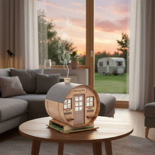 Räuchermännchen „Räucherfass“ (10 cm), farbig gestaltet als kleines Fasshaus mit Tür und Fenstern, rauchend auf einem Tisch in moderner Wohnatmosphäre mit Blick durch ein großes Fenster auf Garten und Campingwagen bei Sonnenuntergang dekoriert.