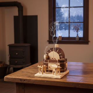 Räuchermännchen „Backofen“ (15 cm), naturbelassen gestaltet als kleiner Holzbackofen mit Backutensilien und Teigwerkzeug, rauchend auf einem Tisch in gemütlicher Wohnatmosphäre vor Ofen und winterlicher Fensterkulisse dekoriert.