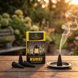 XL-Räucherkerzen „Ruhe!“ (4 Stück), schwarze Duftkegel neben Verpackung mit Aufschrift „RUHE!“. Eine Räucherkerze brennt auf einem kleinen Teller und verströmt Rauch, arrangiert auf einem Holztisch im Garten bei warmem Abendlicht.