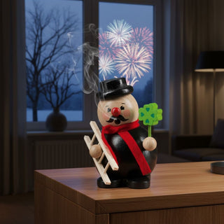 Räuchermännchen „Glücksbringer“ (12 cm), farbig gestaltet mit schwarzem Hut, rotem Schal, Leiter und Kleeblatt in der Hand, rauchend auf einem Tisch vor Fenster mit Feuerwerks‑ und Winterkulisse dekoriert.