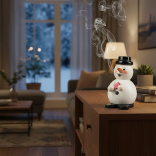 Räuchermännchen „Schneemann mit Herz“ (12 cm), farbig gestaltet mit schwarzem Hut, Karottennase und Herz in der Hand, rauchend auf einem Tisch in gemütlicher Wohnatmosphäre vor winterlicher Fensterkulisse dekoriert.