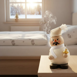 Räuchermännchen „Schlafmütze“ (17 cm), farbig gestaltet mit Zipfelmütze, Schal und Kerzenhalter, rauchend auf einem Nachttisch vor gemütlicher Schlafzimmerszene mit Fensterblick im Winter.