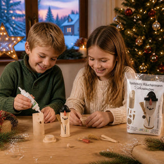 Bastelsatz Räuchermännchen „Schneemann“ aus naturbelassenem Holz, 12 cm. Auf einem Holztisch werden die einzelnen Holzteile eines Räuchermännchens zusammengebaut. Zu sehen sind das bereits teilweise montierte Schneemann-Räuchermännchen, Holzleim, Zubehörteile sowie die Produktverpackung des Bastelsatzes. Im Hintergrund sorgen ein geschmückter Weihnachtsbaum, ein beleuchteter Stern und eine winterliche Fensterkulisse für eine festliche Weihnachtsatmosphäre. Ideal für kreativen Bastelspaß in der Adventszeit.
