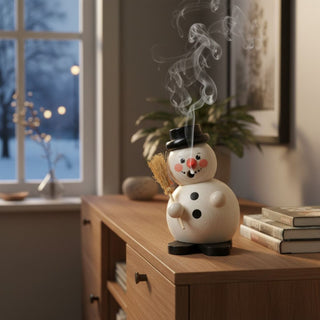 Räuchermännchen „Schneemann mit Besen“ (12 cm), farbig gestaltet mit schwarzem Hut, Karottennase und Besen in der Hand, rauchend auf einem Tisch in gemütlicher Wohnatmosphäre vor winterlicher Fensterkulisse dekoriert.