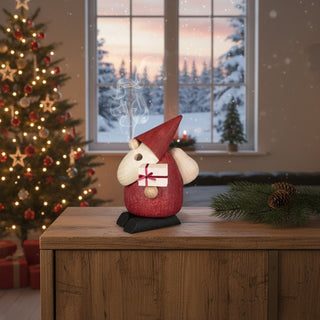 Räuchermännchen „Weihnachtsmann‑Bescherung“ (10 cm), farbig gestaltet mit Geschenk in der Hand, rauchend auf einem Tisch vor festlich geschmücktem Weihnachtsbaum und winterlicher Fensterkulisse dekoriert.