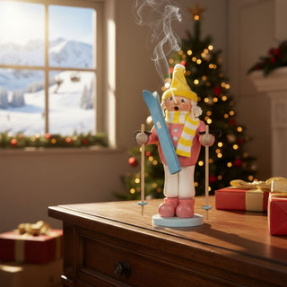 Räuchermännchen „Mädchen mit Ski“ (23 cm), farbig gestaltet mit Mütze, Schal, Skiern und Stöcken, rauchend auf einer Kommode in festlich geschmückter Wohnatmosphäre mit Weihnachtsbaum und winterlicher Berglandschaft im Hintergrund dekoriert.