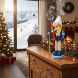 Räuchermännchen „Junge mit Ski“ (23 cm), farbig gestaltet mit Mütze, Schal, Skiern und Stöcken, rauchend auf einer Kommode vor winterlicher Berglandschaft mit Skilift, Weihnachtsbaum und gemütlicher Wohnatmosphäre dekoriert.
