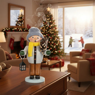 Räuchermännchen „Winterkind“ (21 cm), farbig gestaltet mit Mütze, Schal, Laterne und Bündel Holz, rauchend auf einer Kommode in festlich geschmückter Wohnatmosphäre mit Weihnachtsbaum und winterlicher Schneelandschaft im Hintergrund dekoriert.
