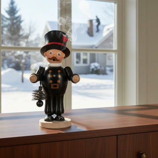 Räuchermännchen „Schornsteinfeger“ (23 cm), farbig gestaltet mit schwarzer Uniform, Zylinder und Besen, rauchend auf einer Kommode vor Fenster mit winterlicher Schneelandschaft dekoriert.