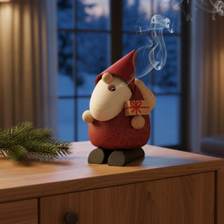 Räuchermännchen „Weihnachtsmann‑Bescherung“ (24 cm), farbig gestaltet mit Geschenk in der Hand, rauchend auf einem Tisch vor gemütlicher Winterkulisse dekoriert.