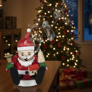 Räuchermännchen Mini „Weihnachtsmann“ (8 cm), farbig gestaltet mit roter Mütze, Glöckchen und kleinem Baum, rauchend auf einem Tisch vor festlich geschmücktem Weihnachtsbaum und warm beleuchteter Wohnkulisse dekoriert.
