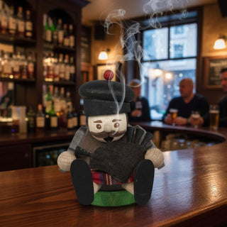 Räuchermännchen Mini „Schotte“ (8 cm), farbig gestaltet mit traditioneller Mütze und Dudelsack, rauchend auf einem Tresen in gemütlicher Bar‑Atmosphäre mit beleuchteten Regalen im Hintergrund dekoriert.