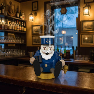 Räuchermännchen Mini „Kapitän“ (8 cm), farbig gestaltet mit blauer Uniform, Anker-Mütze und Fernrohr, rauchend auf einem Tresen in maritimer Bar‑Atmosphäre mit beleuchteten Regalen im Hintergrund dekoriert.